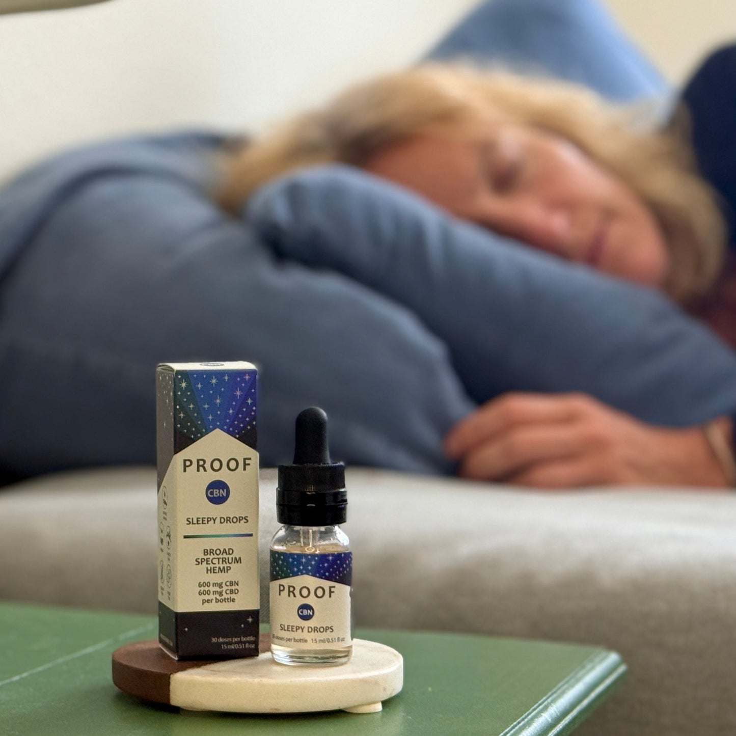 Proof Remedy CBN Sleepy Drops bottle and packaging on nightstand with woman resting on couch in the background.