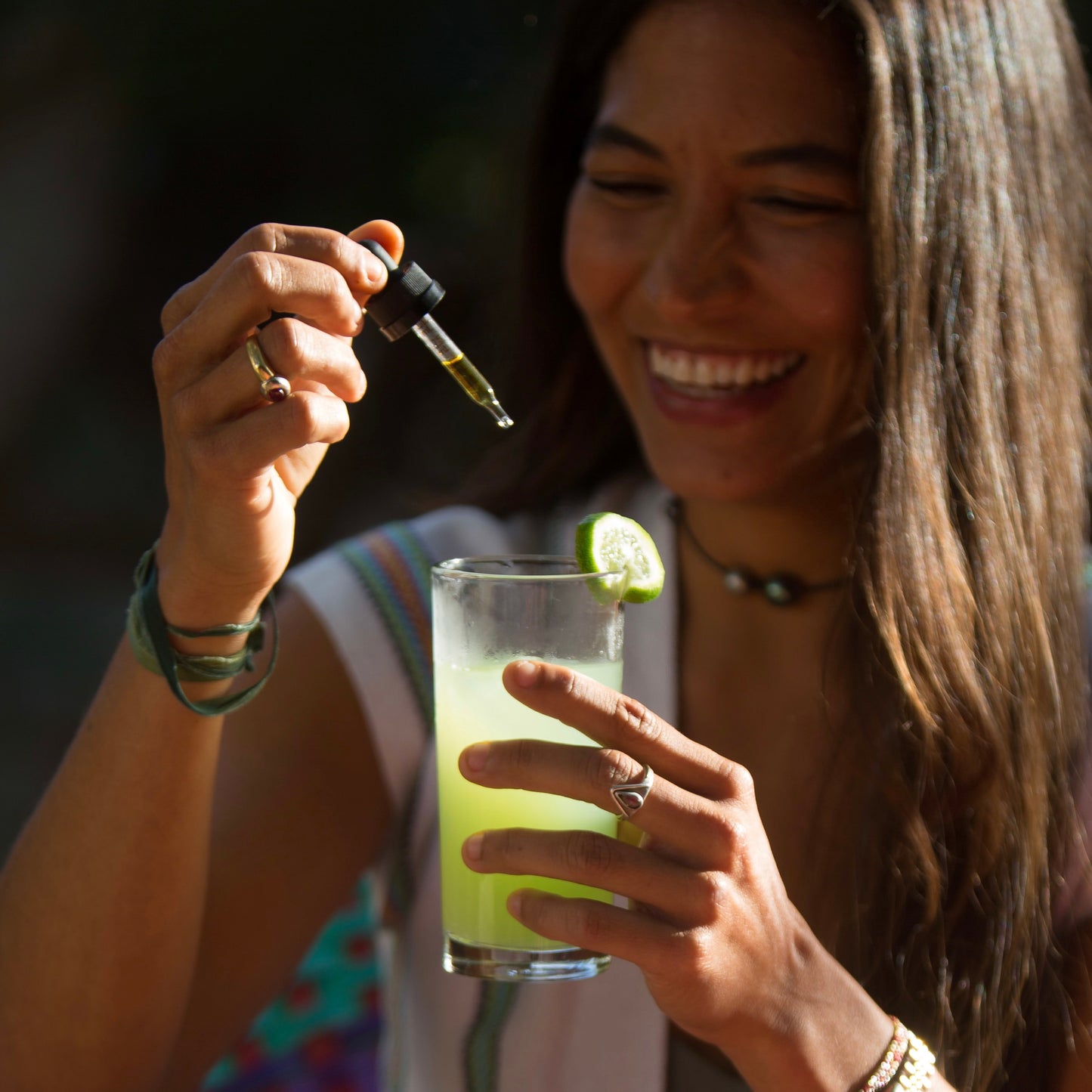 Smiling woman adding Proof Remedy CBD tincture drops into a lime drink, showing plant-based wellness and relaxation.