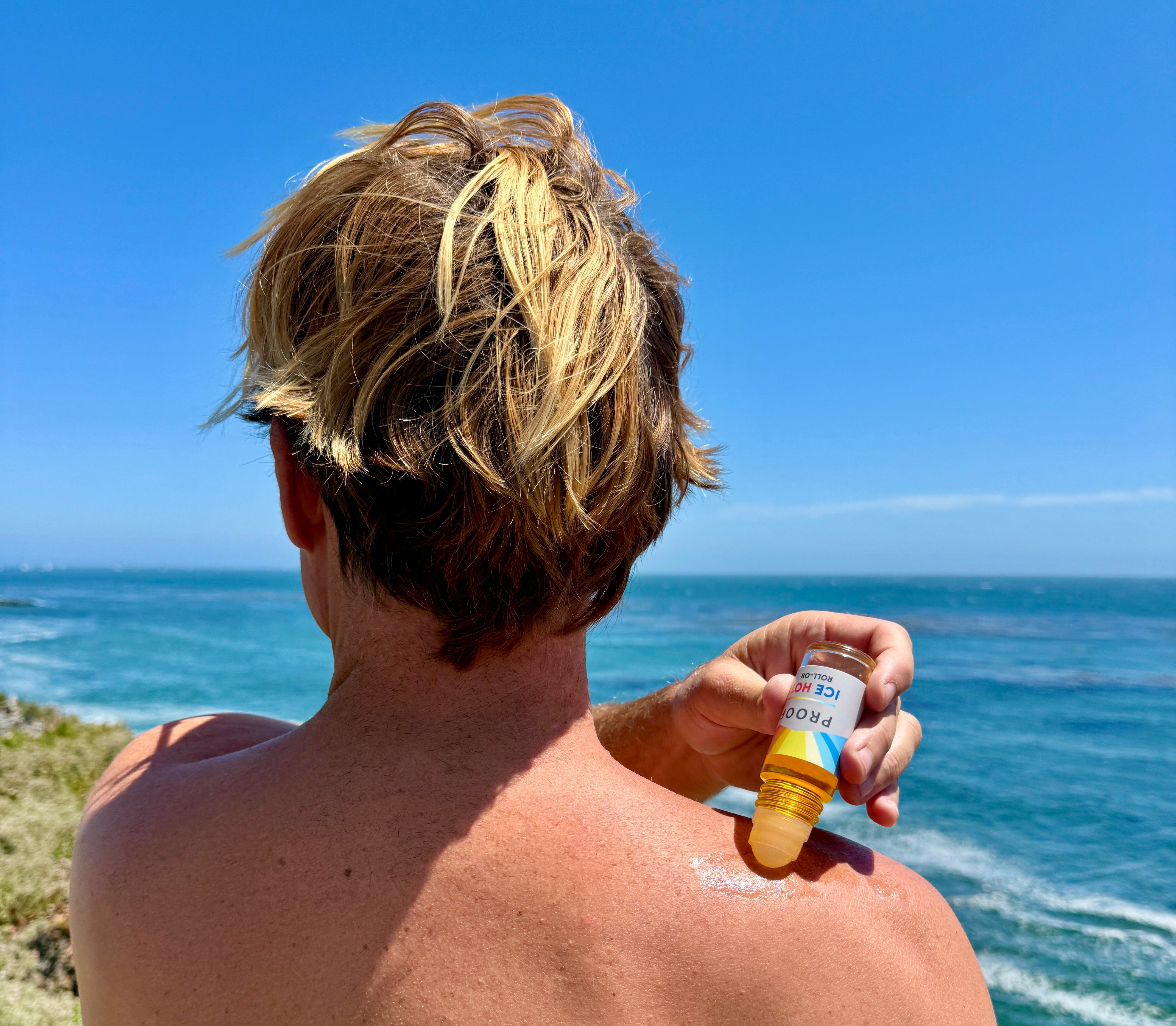 Man applying Proof Remedy Ice Hot Roll-On to shoulder outdoors by the ocean for natural muscle and joint relief.