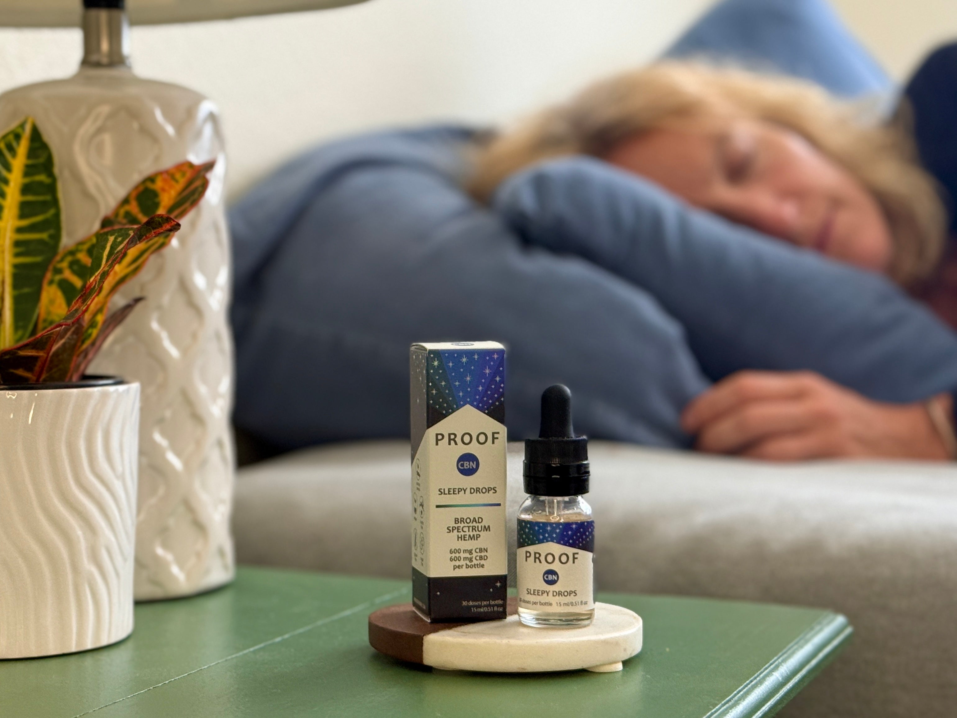 Proof Remedy CBN Sleepy Drops bottle and packaging on nightstand with woman resting on couch in the background.