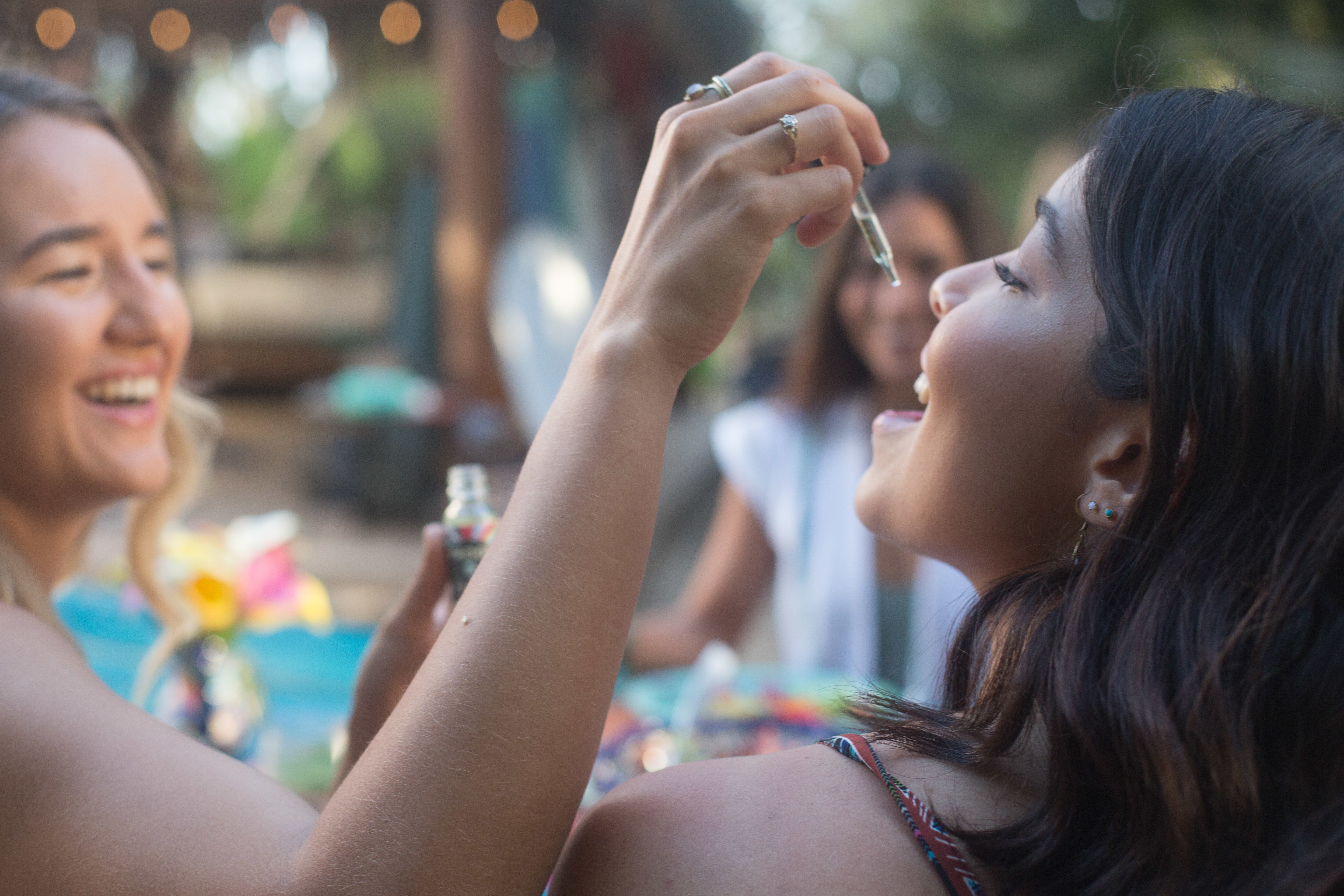 Two women smiling as one uses a dropper to give Proof hemp tincture outdoors at a gathering.