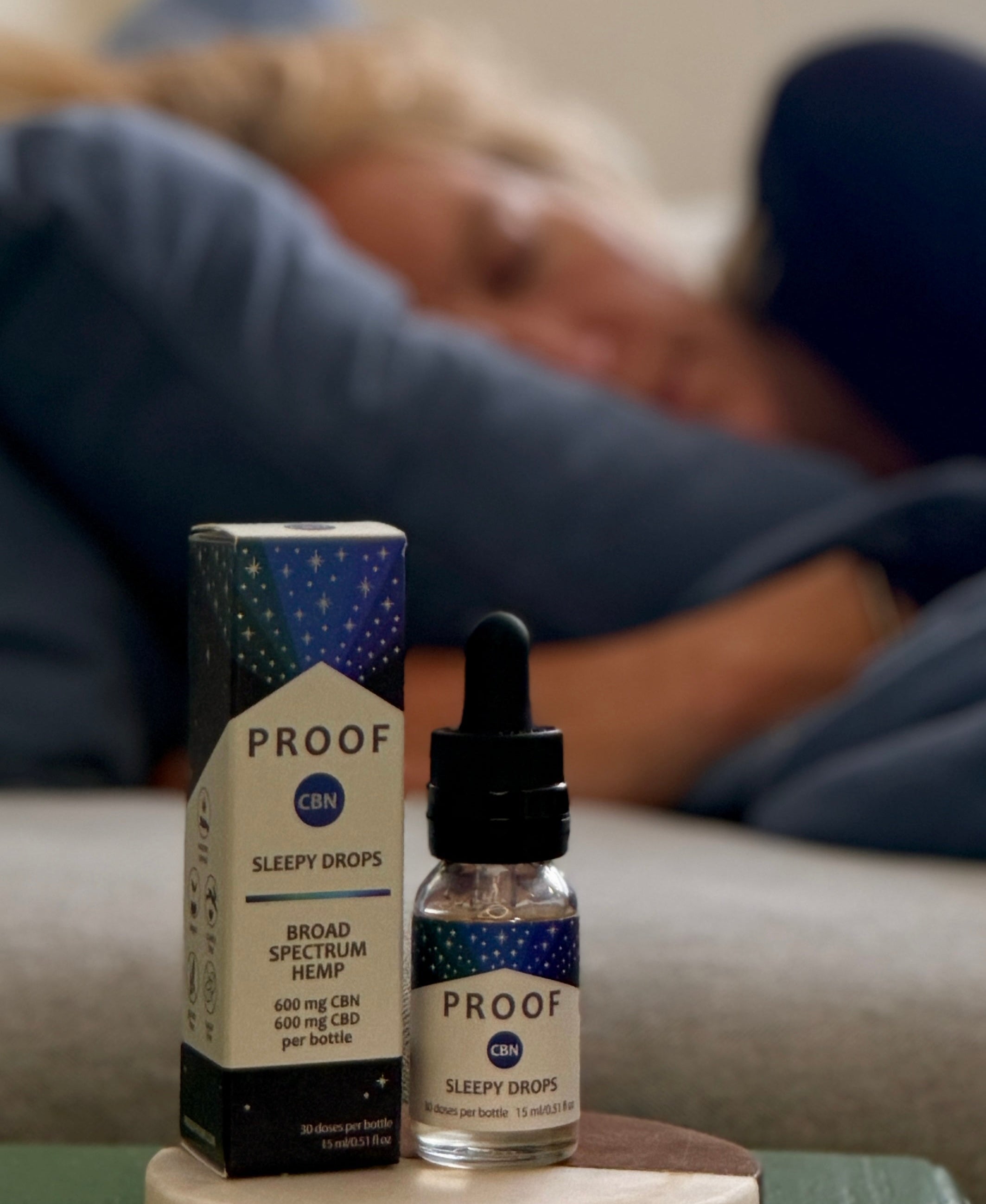 Proof Remedy CBN Sleepy Drops bottle and packaging on nightstand with woman resting on couch in the background.
